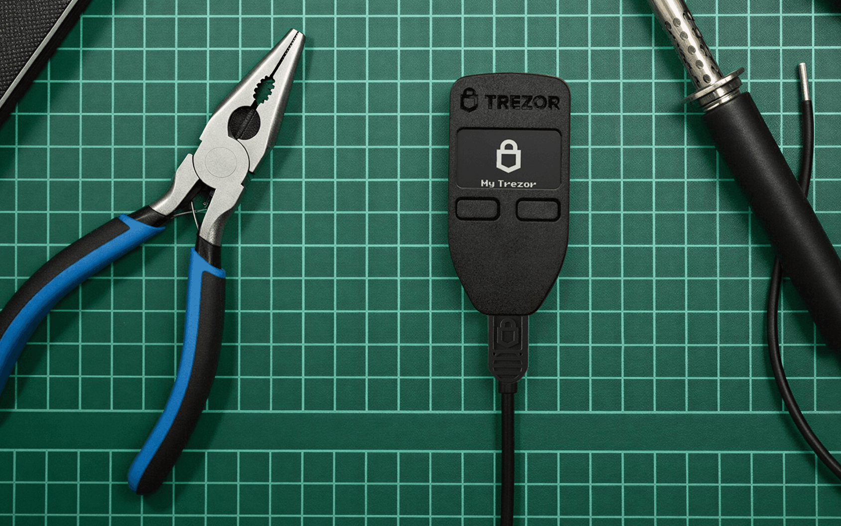 Trezor hardware wallet with pliers and a soldering iron on a green gridded cutting mat.