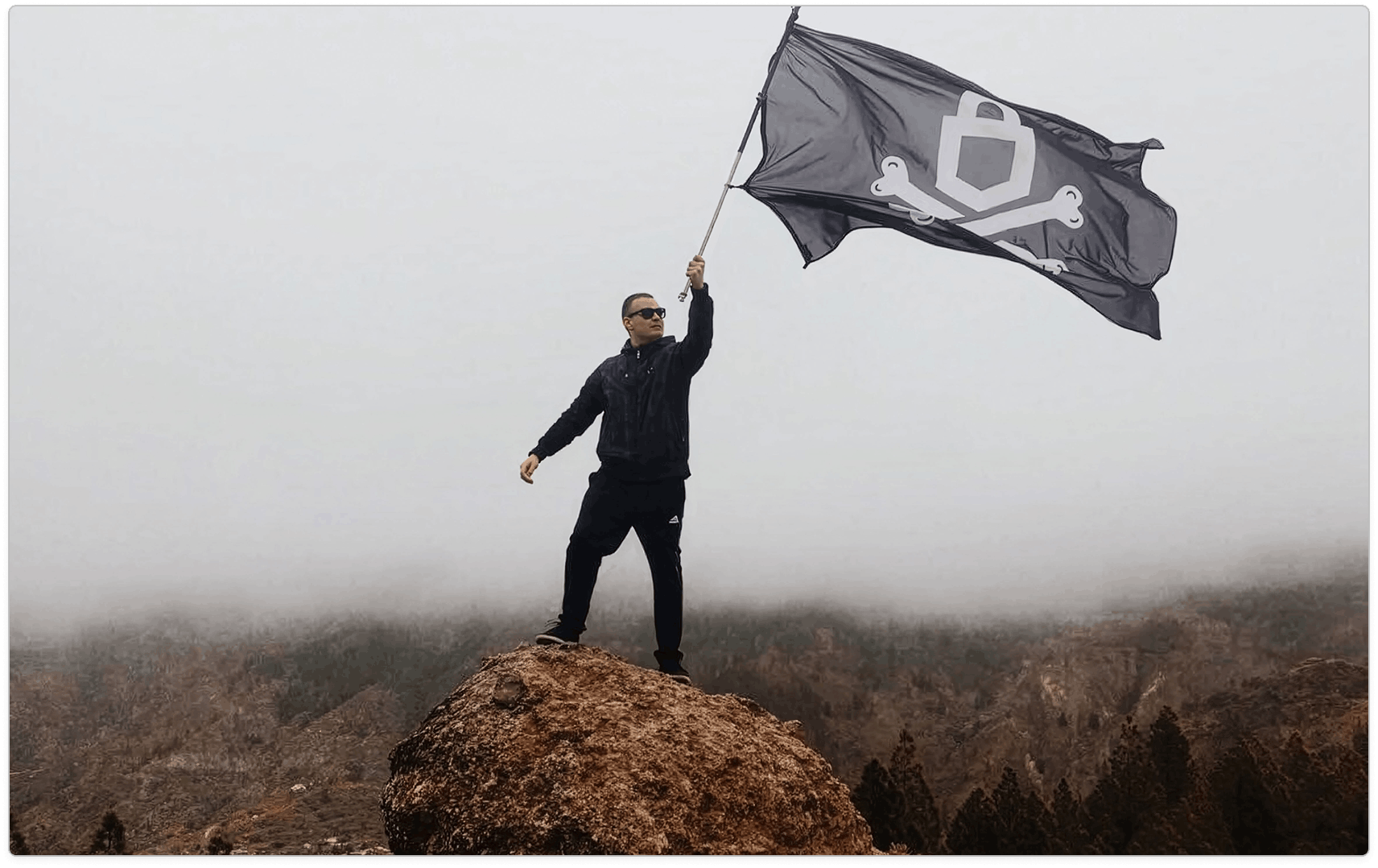 Man in black standing on a rocky outcrop and holding a large dark emblem flag above a foggy forest.