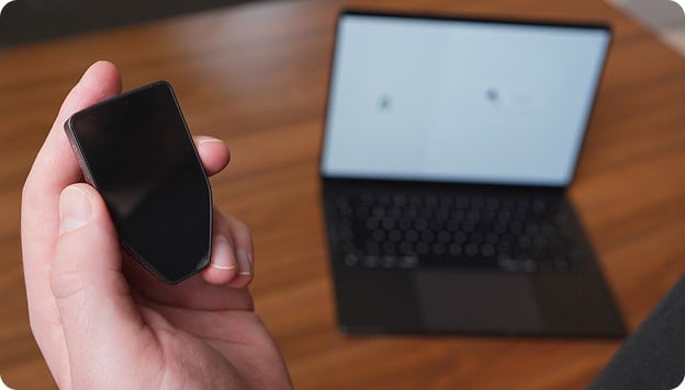 Hand holding a small black electronic device in front of an open laptop on a wooden desk.
