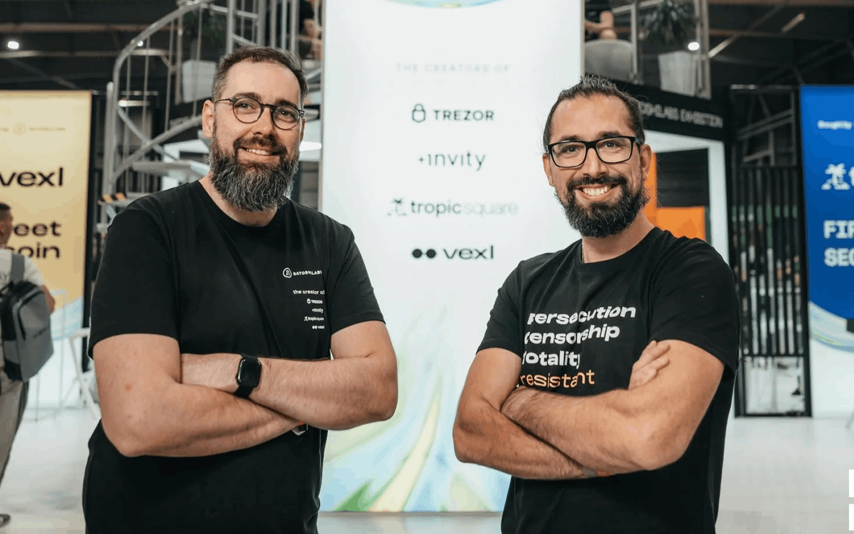 Two bearded men in black T-shirts pose with crossed arms at an indoor technology event.